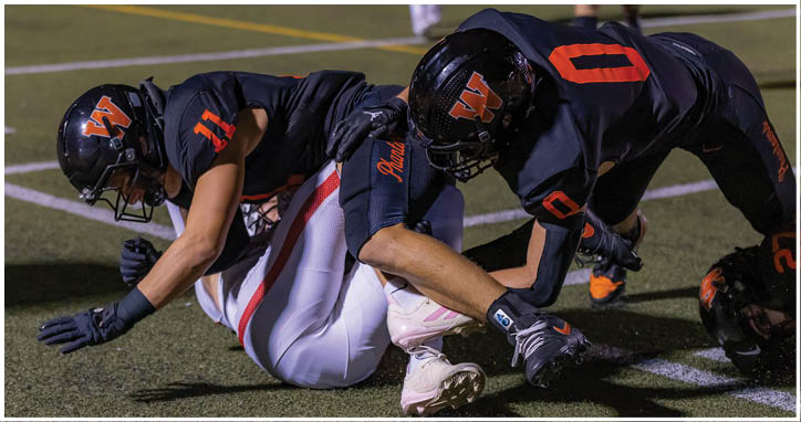 Two people in black football uniforms with orange accents are engaged in a tackle on a green field under stadium lighting. Description generated by AI