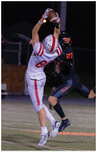Two people in football uniforms are jumping and reaching to catch a football during a nighttime game on a field. Description generated by AI
