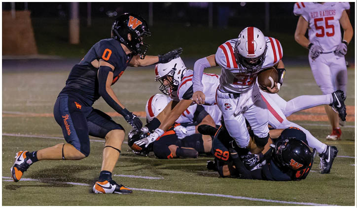 A group of people in football uniforms are actively engaged in a play on a field, with one person running while holding the ball and others attempting to tackle or block. Description generated by AI