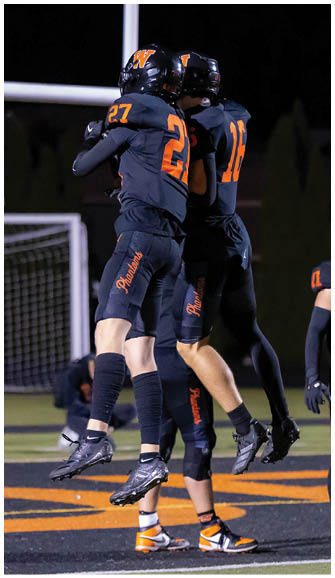 Two people in dark football uniforms with orange numbers are jumping and celebrating on a field at night. Description generated by AI