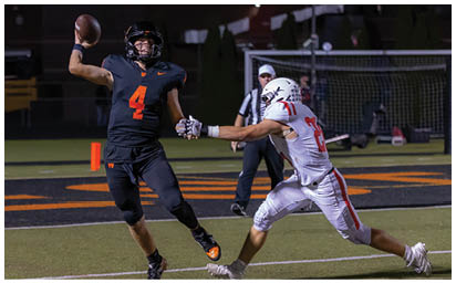 A person in a black football uniform is holding a football and evading a person in a white football uniform who is reaching out to tackle them on a football field at night. Description generated by AI