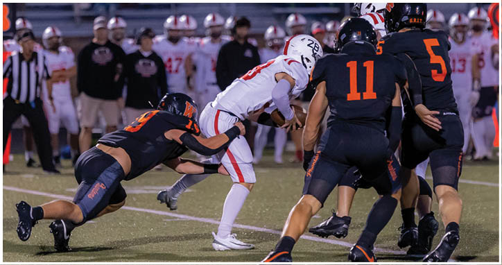 Several people in football uniforms are actively engaged in a play on a field, with one person in white being tackled by others in dark uniforms. Description generated by AI