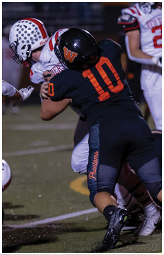 Two people in football uniforms are engaged in a tackle on a field while a third person in a white uniform stands nearby. Description generated by AI