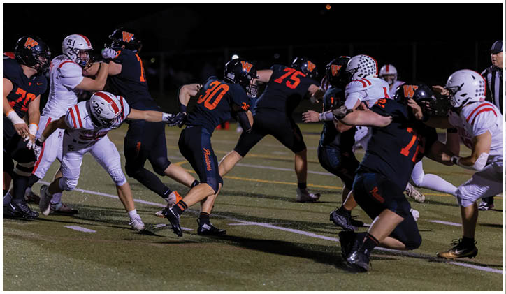 People in football uniforms are actively engaged in a game on a field under stadium lights, with some players tackling and others running or blocking. Description generated by AI