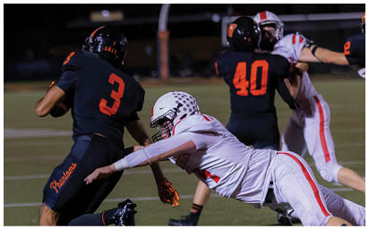 A person in a black football uniform with the number 3 is running while another person in a white football uniform is diving to tackle them on a lit football field at night. Description generated by AI