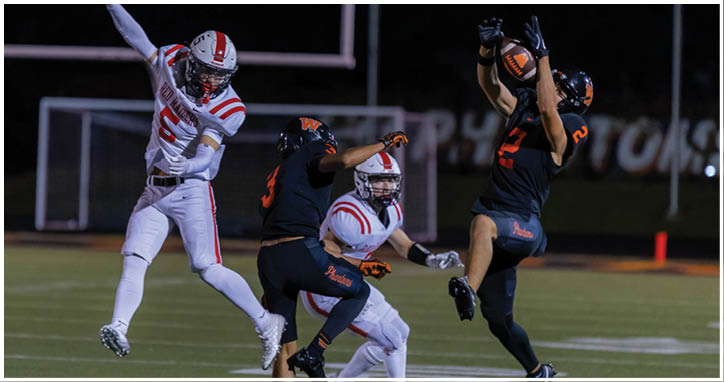 A group of people in football uniforms are actively engaged in a play on a field at night, with one person in white throwing a ball and others in dark uniforms attempting to catch or block it. Description generated by AI
