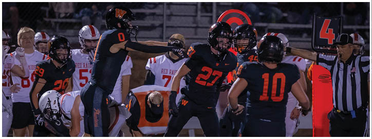 Several people in football uniforms are gathered on a field near the sideline with a referee holding a marker in the background. Description generated by AI