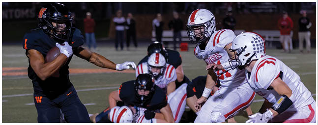People in football uniforms are actively engaged in a play on a lit field during a game. Description generated by AI