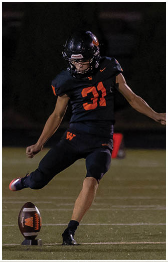 A person wearing a black football uniform with the number 31 is in the motion of kicking a football on a field at night. Description generated by AI