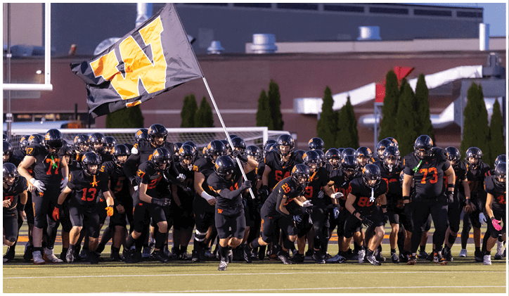 A large group of people in black football uniforms with orange numbers are running onto a field, with one person holding a flag featuring a large yellow “W". Description generated by AI