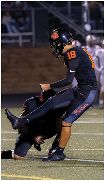 A person in a black football uniform with the number 18 is kicking a football held by another person kneeling on the field during a nighttime game. Description generated by AI