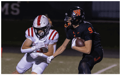 Two people in football uniforms are engaged in a play with one person holding the ball and the other attempting to block or tackle. Description generated by AI