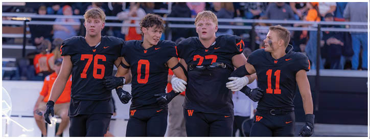 Four adults in black and orange football uniforms stand arm in arm on a field with a crowd in the background. Description generated by AI