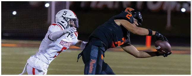 A person in dark sportswear is reaching out to catch a football while another person in white sportswear is attempting to tackle them on a football field at night. Description generated by AI