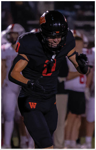A person in black football gear is running on a field while a referee in a striped shirt raises one hand and people in white football uniforms stand in the background. Description generated by AI