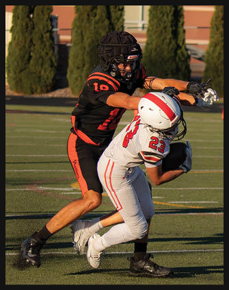 Two people in football uniforms are engaged in a play on a football field, with one person in white and red attempting to catch or hold the ball while the other in black and orange reaches toward them. Description generated by AI