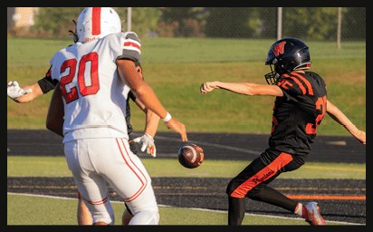 A person in a black and orange football uniform is kicking a football while two people in white and red uniforms are nearby on a football field. Description generated by AI