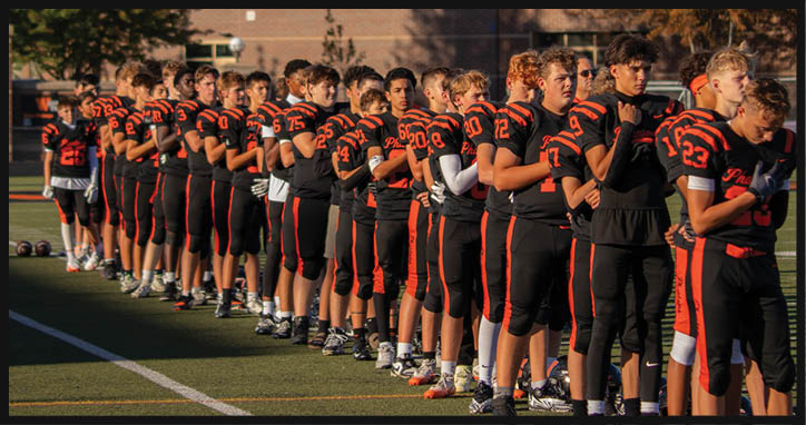 A line of people in black and orange football uniforms stand side by side on a field with their hands over their hearts. Description generated by AI
