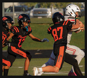 Two people in black and orange football uniforms are attempting to tackle a person in a white football uniform holding a football on a grassy field. Description generated by AI