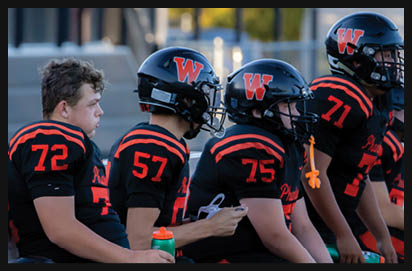 Four people in black and orange football uniforms sit on a bench with helmets nearby, one holding a water bottle and another holding a phone. Description generated by AI