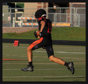 A person wearing a black and orange football uniform is running on a football field while holding a football. Description generated by AI