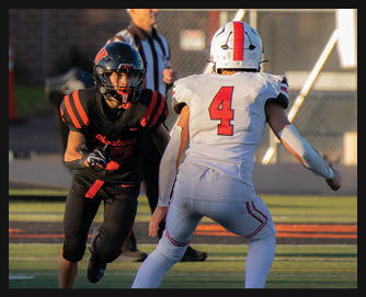 Two people in football uniforms are facing each other on a field with one person in black and red preparing to move and the other in white and red standing ready. Description generated by AI