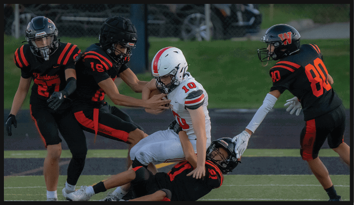 Three people in football uniforms are engaged in a play on a field, with one person in white being tackled by two people in black and orange. Description generated by AI