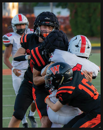 Two people in football uniforms are engaged in a tackle on a field with one wearing a black and orange jersey and the other in a white jersey with a red stripe on the helmet. Description generated by AI