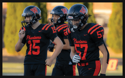 Three people wearing black and red football uniforms with helmets stand on a field, with two of them visible in the foreground and one slightly blurred in the background. Description generated by AI
