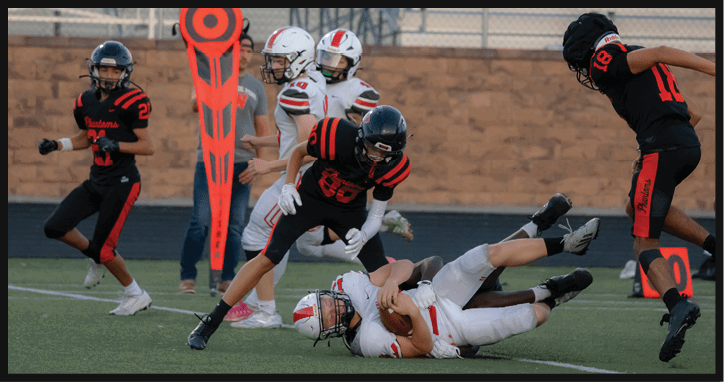 People in football uniforms are playing on a field with one person in white being tackled to the ground by a person in black and red, while others stand nearby and a down marker is visible. Description generated by AI