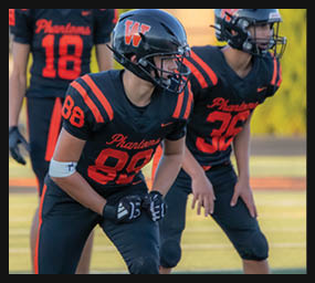 Three people in black and orange football uniforms and helmets are positioned on a field, appearing ready for action. Description generated by AI
