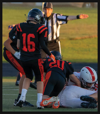 Two people in black and red football uniforms with helmets are on a field, one standing with the number 16 on the back and the other kneeling with the number 20, while a referee in a black and white striped shirt gestures in the background. Description generated by AI