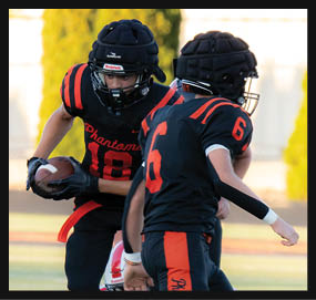 Two people wearing black and orange football uniforms and helmets are on a field with one person holding a football. Description generated by AI