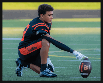 A person in a black and red football uniform is kneeling on a field while holding a football upright on the ground. Description generated by AI