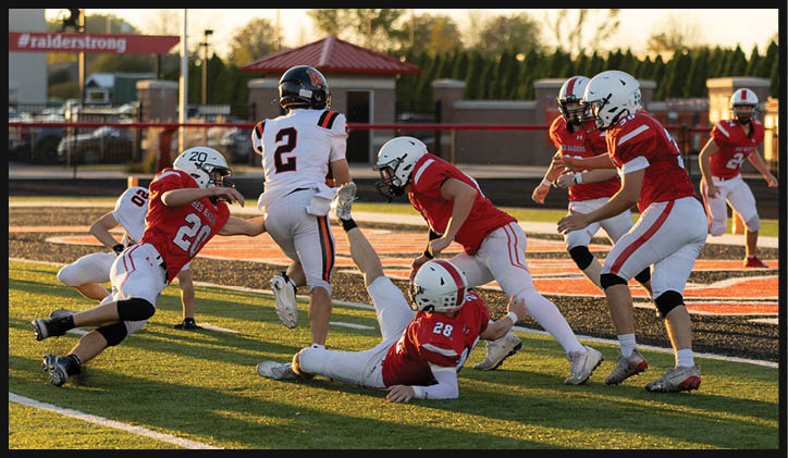 Several people wearing football uniforms are actively engaged in a play on a sunlit field with one person in white running while others in red attempt to tackle or block. Description generated by AI