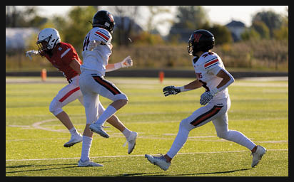 Two people in football uniforms are actively engaged on a sunlit field with one person in a red jersey and white helmet and the other in a white jersey and black helmet as a football flies through the air nearby. Description generated by AI