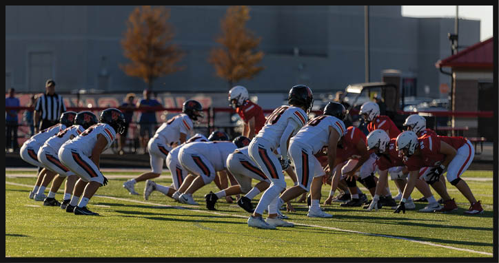 Two teams of people in football uniforms are lined up facing each other on a sunlit field preparing for a play. Description generated by AI