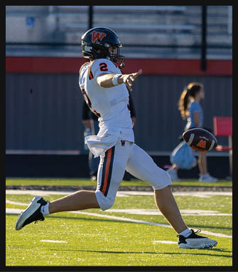 A person in a white football uniform with a black helmet is kicking a football on a sunlit field with a blurred background of people and bleachers. Description generated by AI
