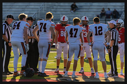 People in football uniforms and referees stand together on a field during a game with empty bleachers in the background. Description generated by AI