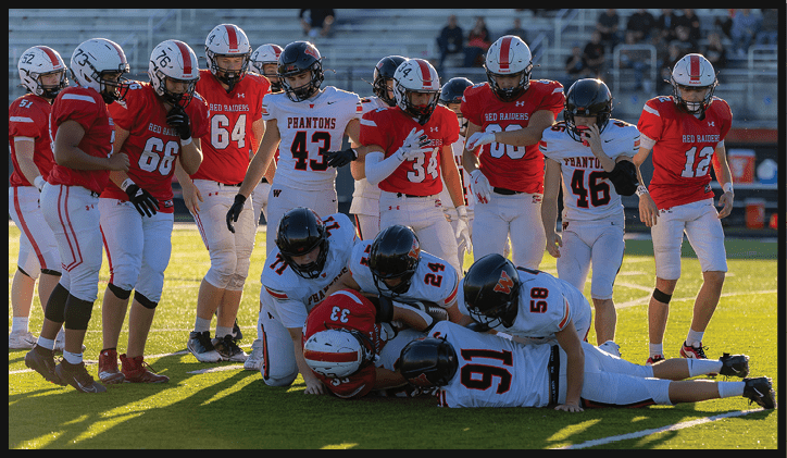 Several people in football uniforms are on a field with some players in red standing and others in white lying on the ground in a pile. Description generated by AI