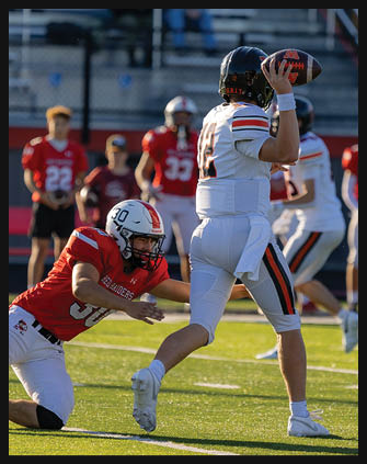 A person in a white football uniform is preparing to throw a football while another person in a red uniform is attempting to tackle them on a grassy field with other players and spectators in the background. Description generated by AI