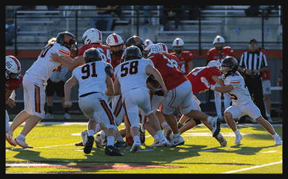 A group of people wearing football uniforms are engaged in a play on a sunlit field with a referee and empty bleachers in the background. Description generated by AI