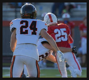 Two people in football uniforms with helmets are on a field, one wearing a white jersey with the number 24 and the other in a red jersey with the number 25. Description generated by AI