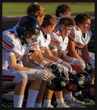 A group of people in white sports uniforms with red and black accents sit closely together on a bench holding black helmets with a red “W" logo, appearing focused and attentive. Description generated by AI
