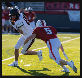 Two people in football uniforms are on a field with one person in a red jersey lunging to tackle the other person in a white jersey holding a football. Description generated by AI