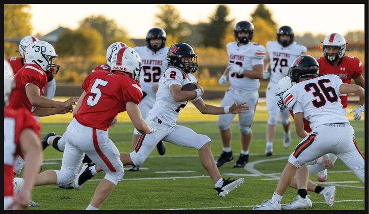 People in football uniforms are actively playing on a field with one person running with the ball while others attempt to block or tackle. Description generated by AI