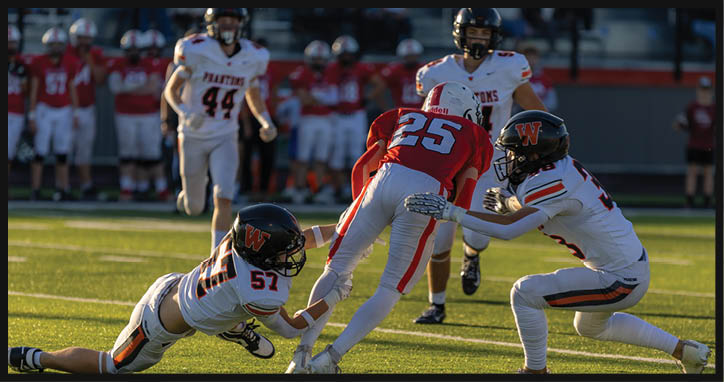A group of people in football uniforms are actively playing on a field with two players in white uniforms attempting to tackle a player in a red uniform running with the ball. Description generated by AI