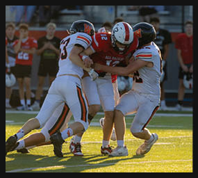 Two people in white sports uniforms tackle a person in a red sports uniform during a football game on a sunlit field. Description generated by AI