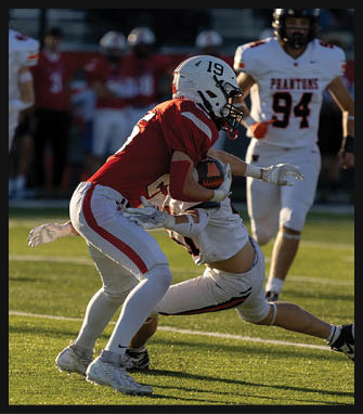 A person in a red football uniform with the number 19 is holding a football while being tackled by a person in a white uniform on a grassy field with another player in white standing nearby. Description generated by AI