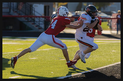 A person in a white and black football uniform is running with the ball while another person in a red and white uniform attempts to tackle them on a sunlit football field. Description generated by AI